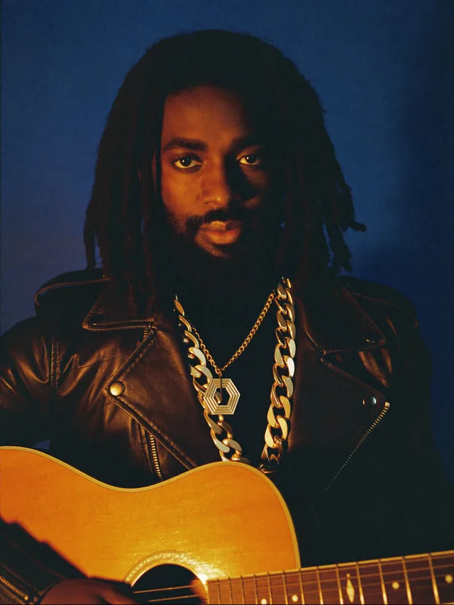 The RumbaMi guitarist, acoustic guitar held against him, leather jacket and gold chain, against a midnight-blue backdrop.